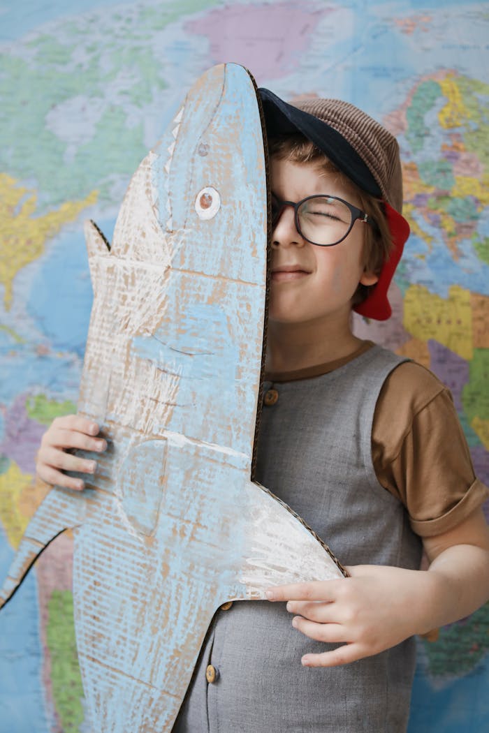 A young boy in glasses and a hat plays with a cardboard shark in front of a world map, showcasing creativity.